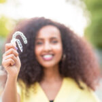 Blurred Black woman in a yellow blouse smiles while holding her Invisalign clear aligners in front of her outside