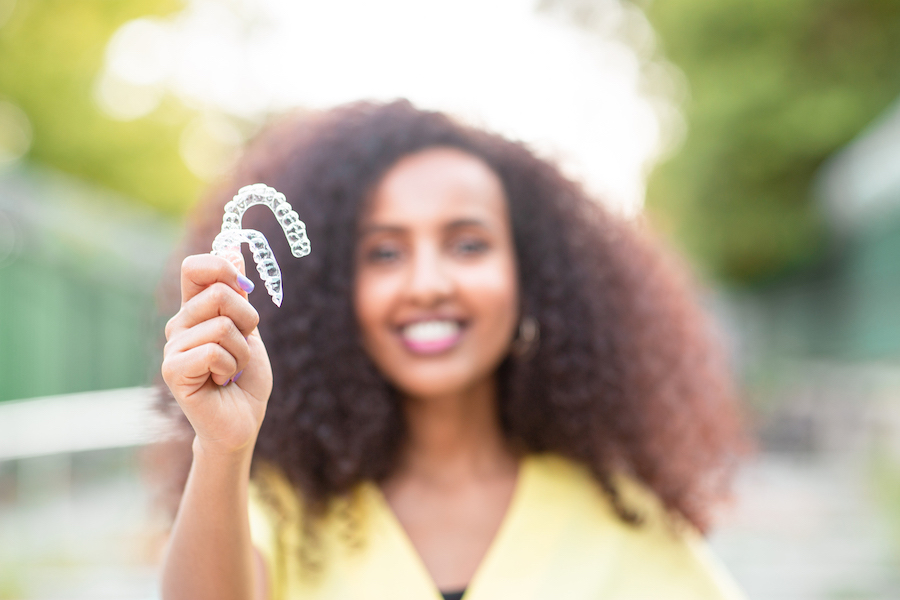 Blurred Black woman in a yellow blouse smiles while holding her Invisalign clear aligners in front of her outside